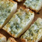 Several pieces of garlic bread with melted cheese and herbs on top are arranged on a wooden surface. The bread appears to be sliced into rectangular pieces, displaying a crispy texture on the edges and a soft, fluffy interior.