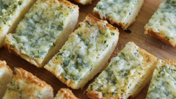 Several pieces of garlic bread with melted cheese and herbs on top are arranged on a wooden surface. The bread appears to be sliced into rectangular pieces, displaying a crispy texture on the edges and a soft, fluffy interior.