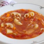 A white bowl filled with minestrone soup, featuring ingredients such as elbow pasta, diced carrots, tomatoes, and a broth, sits on a white surface. A white and red patterned cloth is partially visible in the background.