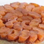 A round caramelized apple tart is displayed on a clear glass plate. The tart features neatly arranged, overlapping apple slices, golden and glossy from the caramel glaze on top. The crust appears slightly visible at the edges, hinting at a flaky pastry base.