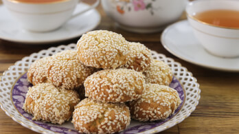 A plate of sesame-covered cookies is neatly arranged on a decorative purple and white plate. Behind the plate, there are two white teacups filled with tea and a floral-patterned teapot, all set on a wooden table.