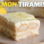 A slice of lemon tiramisu sits on a plate, garnished with grated lemon zest. The background features whole lemons and a blurred view of the full tiramisu dessert. The words "LEMON TIRAMISU" are prominently displayed at the top of the image.