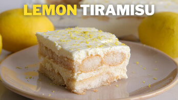 A slice of lemon tiramisu sits on a plate, garnished with grated lemon zest. The background features whole lemons and a blurred view of the full tiramisu dessert. The words "LEMON TIRAMISU" are prominently displayed at the top of the image.