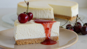 A slice of cheesecake topped with red cherry sauce and two fresh cherries on a beige plate. The creamy cheesecake has a golden brown crust. In the background, there's a whole cheesecake with a slice missing, and more cherries are scattered beside the plate.