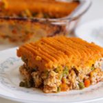 A slice of shepherd's pie with a layer of mashed sweet potatoes on top and a filling of ground meat, peas, and carrots on a white plate. The remainder of the pie is visible in a glass baking dish in the background.