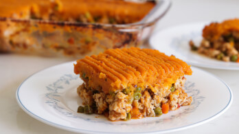 A slice of shepherd's pie with a layer of mashed sweet potatoes on top and a filling of ground meat, peas, and carrots on a white plate. The remainder of the pie is visible in a glass baking dish in the background.