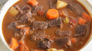 A bowl of hearty beef stew filled with chunks of tender beef, carrots, potatoes, and pieces of tomato, all immersed in a rich, brown broth.