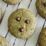 Six cookies with a green hue, possibly made with matcha or another green ingredient, are cooling on a wire rack. Each cookie is decorated with three chocolate chips arranged in a triangle pattern.