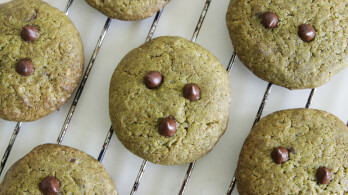 Six cookies with a green hue, possibly made with matcha or another green ingredient, are cooling on a wire rack. Each cookie is decorated with three chocolate chips arranged in a triangle pattern.