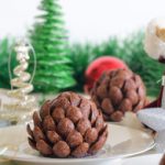 A festive scene featuring chocolate pine cone desserts on a white plate, surrounded by Christmas ornaments and decorations. There is a Santa figurine holding a silver star ornament, with green tinsel and Christmas trees in the background.