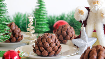 A festive scene featuring chocolate pine cone desserts on a white plate, surrounded by Christmas ornaments and decorations. There is a Santa figurine holding a silver star ornament, with green tinsel and Christmas trees in the background.