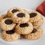 A plate of thumbprint cookies filled with chocolate sits on a white surface. Next to the plate is a set of gold utensils and a red cloth. The cookies are dusted with crushed nuts, and a single cookie rests on the red cloth.