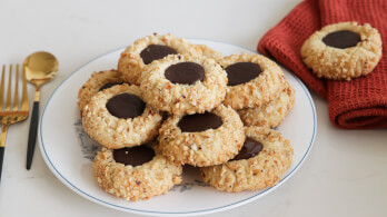 A plate of thumbprint cookies filled with chocolate sits on a white surface. Next to the plate is a set of gold utensils and a red cloth. The cookies are dusted with crushed nuts, and a single cookie rests on the red cloth.