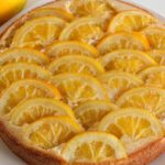 A round lemon cake on a white plate is topped with neatly arranged semi-circular slices of lemon. Two whole lemons are placed next to the plate on a light-colored surface.