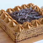 A square coffee-flavored cake topped with rosettes of coffee frosting and a generous amount of chocolate shavings in the center. The cake sits on a wooden board, with a white background.