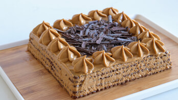 A square coffee-flavored cake topped with rosettes of coffee frosting and a generous amount of chocolate shavings in the center. The cake sits on a wooden board, with a white background.