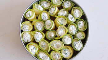 A round baking dish filled with rolled zucchini slices, each containing a creamy white filling with herbs. The zucchini rolls are arranged upright, closely packed together in the dish.