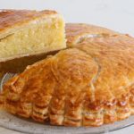 A round Galette des Rois, a traditional French cake with a flaky, golden-brown crust, is displayed on a glass plate. A slice has been cut and is slightly lifted, revealing a dense, almond frangipane filling inside.