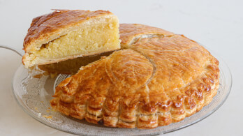 A round Galette des Rois, a traditional French cake with a flaky, golden-brown crust, is displayed on a glass plate. A slice has been cut and is slightly lifted, revealing a dense, almond frangipane filling inside.