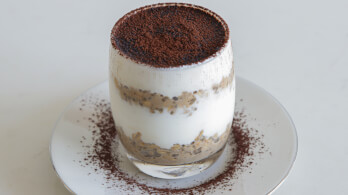 A dessert layered in a transparent glass is placed on a white plate. The dessert has visible layers of creamy white substance and a brown ingredient, possibly cocoa or coffee powder, sprinkled on top. There is also some powdered topping on the plate.