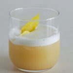 A clear glass filled with a creamy, light beige cocktail topped with a frothy foam. A lemon peel garnish floats on top of the foam. The drink is set against a plain, light grey background.