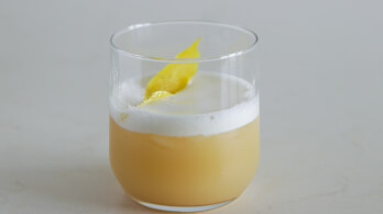 A clear glass filled with a creamy, light beige cocktail topped with a frothy foam. A lemon peel garnish floats on top of the foam. The drink is set against a plain, light grey background.