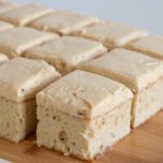 A wooden tray holds a neatly arranged grid of frosted vanilla cake squares. The cake has a dense, moist texture and is topped with a smooth layer of light brown frosting. The background is plain and white, placing full emphasis on the cake.