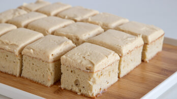 A wooden tray holds a neatly arranged grid of frosted vanilla cake squares. The cake has a dense, moist texture and is topped with a smooth layer of light brown frosting. The background is plain and white, placing full emphasis on the cake.