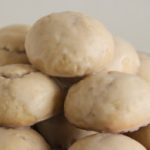 A plate stacked with round, glazed Italian anise cookies. The cookies have a light, golden-brown hue, and the frosting gives them a shiny appearance. The background is plain and neutral, keeping the focus on the cookies.