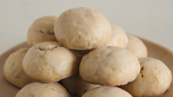 A plate stacked with round, glazed Italian anise cookies. The cookies have a light, golden-brown hue, and the frosting gives them a shiny appearance. The background is plain and neutral, keeping the focus on the cookies.