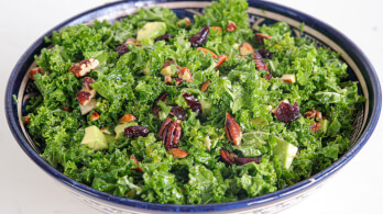 A ceramic bowl filled with a fresh kale salad. The salad is mixed with avocado chunks, dried cranberries, and pecan nuts. The colors of the dark green kale contrast with the vibrant red cranberries and brown pecans, creating an appetizing presentation.