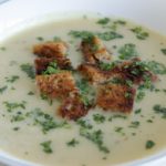 A bowl of creamy soup garnished with fresh chopped parsley and croutons. The soup appears smooth and thick, with the croutons adding texture and color.