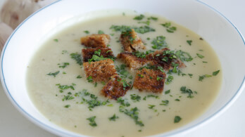 A bowl of creamy soup garnished with fresh chopped parsley and croutons. The soup appears smooth and thick, with the croutons adding texture and color.