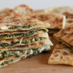 A stack of golden-brown, triangular flatbreads filled with spinach and feta cheese are placed on a wooden surface. Some flatbreads are piled high while others lie flat, showcasing the appetizing, layered filling.