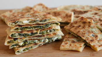 A stack of golden-brown, triangular flatbreads filled with spinach and feta cheese are placed on a wooden surface. Some flatbreads are piled high while others lie flat, showcasing the appetizing, layered filling.