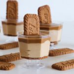 Mini dessert cups in the image have layers of cream and caramel, topped with a Lotus Biscoff cookie. Several more Biscoff cookies are placed around the cups on the white surface. The transparent cups show the distinct layers of the dessert inside.