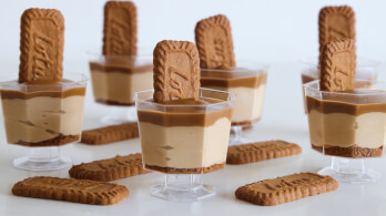 Mini dessert cups in the image have layers of cream and caramel, topped with a Lotus Biscoff cookie. Several more Biscoff cookies are placed around the cups on the white surface. The transparent cups show the distinct layers of the dessert inside.