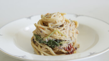 A white plate holding a neatly twirled serving of spaghetti pasta mixed with creamy sauce, green spinach leaves, and bits of red sun-dried tomatoes. The dish is presented in an appetizing manner with a simple, elegant background.