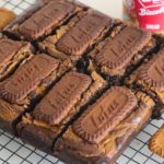 A batch of chocolate brownies topped with Lotus Biscoff cookies, cut into squares, cooling on a wire rack. A jar of Lotus Biscoff spread is visible in the background, next to some additional Biscoff cookies.