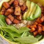 A vibrant bowl of food containing quinoa, diced tofu, sliced avocado, cubed sweet potatoes, and fresh spinach leaves, all arranged in sections. The ingredients are colorful and neatly organized, creating an appetizing and healthy meal.