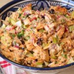 A decorative blue and white bowl filled with a colorful chicken salad featuring shredded chicken, diced vegetables, and dark dried fruits, garnished with fresh herbs. The bowl is placed on a wooden surface next to a red and white checkered cloth with forks and spoons.