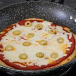 A pan with a freshly cooked pizza topped with melted cheese, tomato sauce, and green olive slices sits on a stovetop. The pizza has a golden-brown crust and is in a speckled black frying pan.