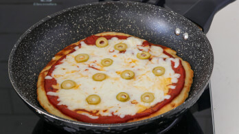 A pan with a freshly cooked pizza topped with melted cheese, tomato sauce, and green olive slices sits on a stovetop. The pizza has a golden-brown crust and is in a speckled black frying pan.