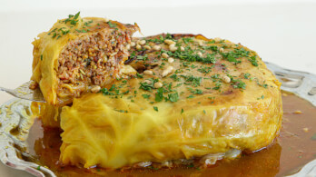 A stuffed cabbage roll topped with pine nuts and chopped herbs, served in a dish with a generous amount of sauce. The cabbage roll is cut to reveal a filling of rice, vegetables, and ground meat.