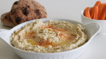 A white dish filled with hummus topped with olive oil and paprika, served with a side of pita bread in the background and a bowl of sliced carrots to the right.