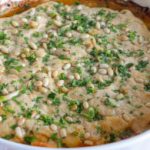 A baked dish in a white round casserole features a golden-brown crust topped with chopped herbs and pine nuts. The edges are slightly crisped, and the dish sits on a red and white checkered cloth.