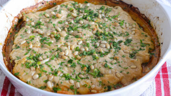 A baked dish in a white round casserole features a golden-brown crust topped with chopped herbs and pine nuts. The edges are slightly crisped, and the dish sits on a red and white checkered cloth.