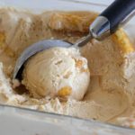 A metal ice cream scoop is scooping out creamy ice cream from a rectangular glass container. The ice cream appears to have churros or pieces of pastry mixed in. The ice cream has a pale, caramel-like color and a smooth texture.