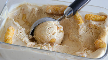 A metal ice cream scoop is scooping out creamy ice cream from a rectangular glass container. The ice cream appears to have churros or pieces of pastry mixed in. The ice cream has a pale, caramel-like color and a smooth texture.