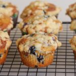 A batch of blueberry muffins topped with crumbly streusel sits on a cooling rack. The muffins are golden brown with visible blueberries peeking through the baked batter. The cooling rack is placed on a white surface.